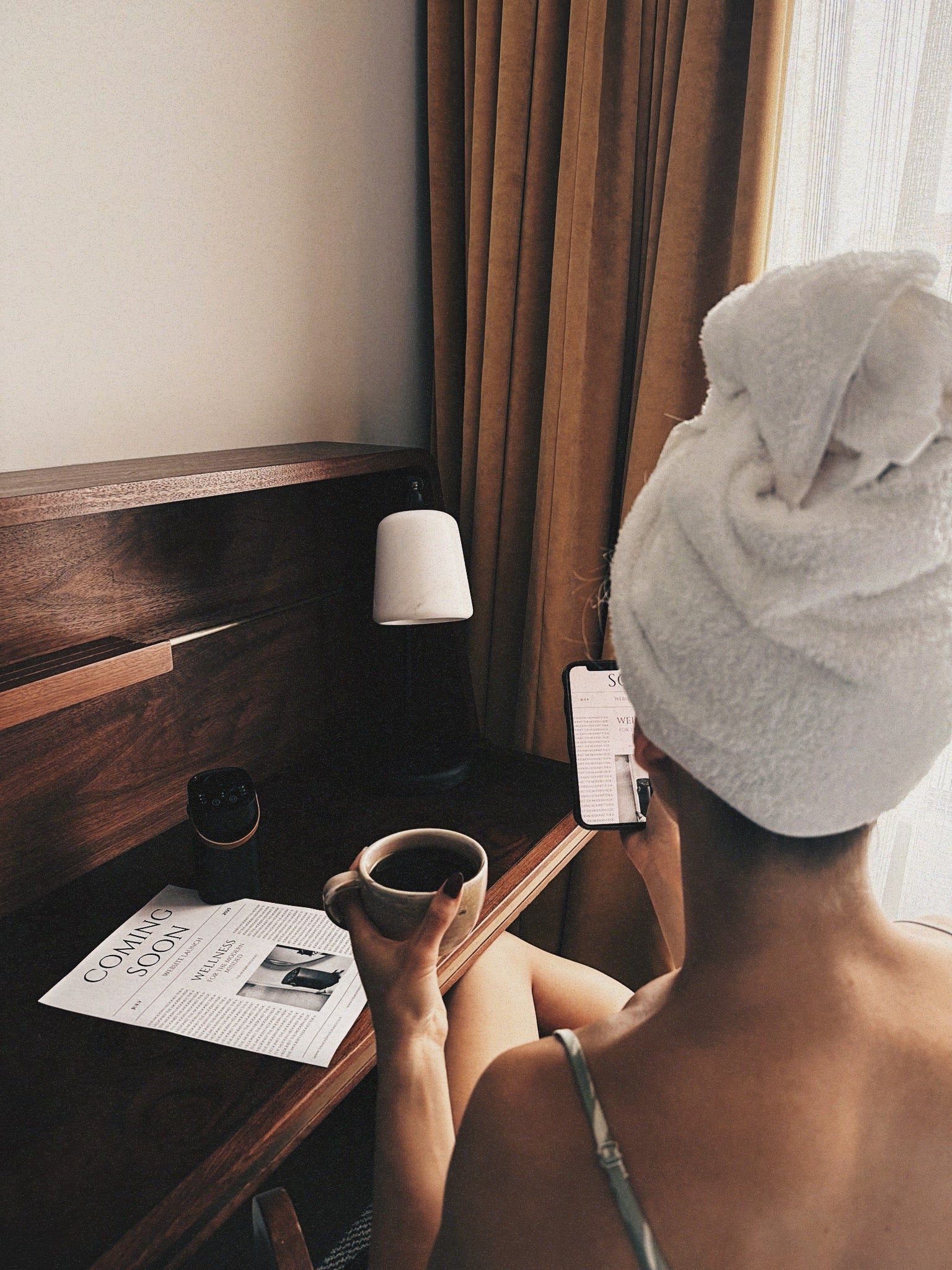 Person with a towel on their head holding a cup, sitting at a desk with a lamp and newspaper.